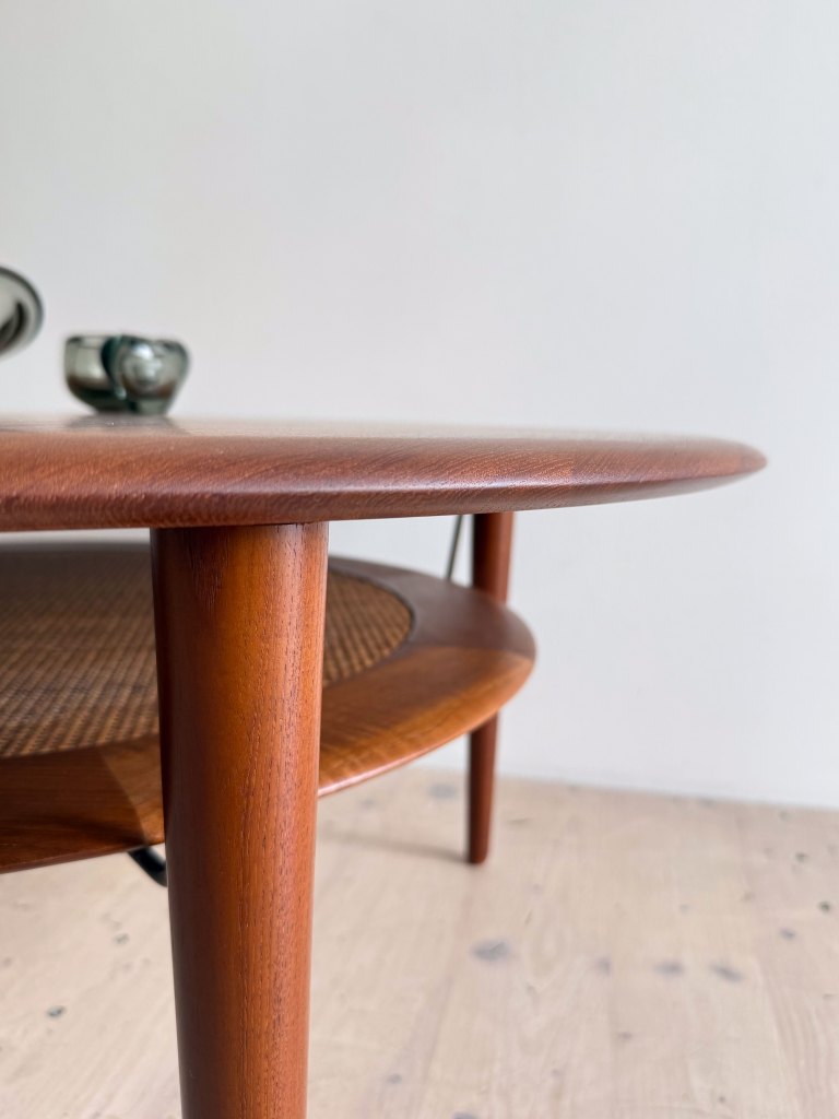 A close-up view of a round wooden coffee table with a smooth finish and tapered legs, featuring a woven lower shelf, placed on a light-colored wooden floor against a plain wall.