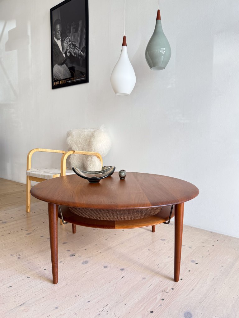 A modern living room scene featuring a round wooden coffee table, a decorative black bowl, and a small stone. Above, three pendant lights hang, and a chair with a white fur throw is visible nearby. A framed black-and-white photograph of a musician is on the wall.