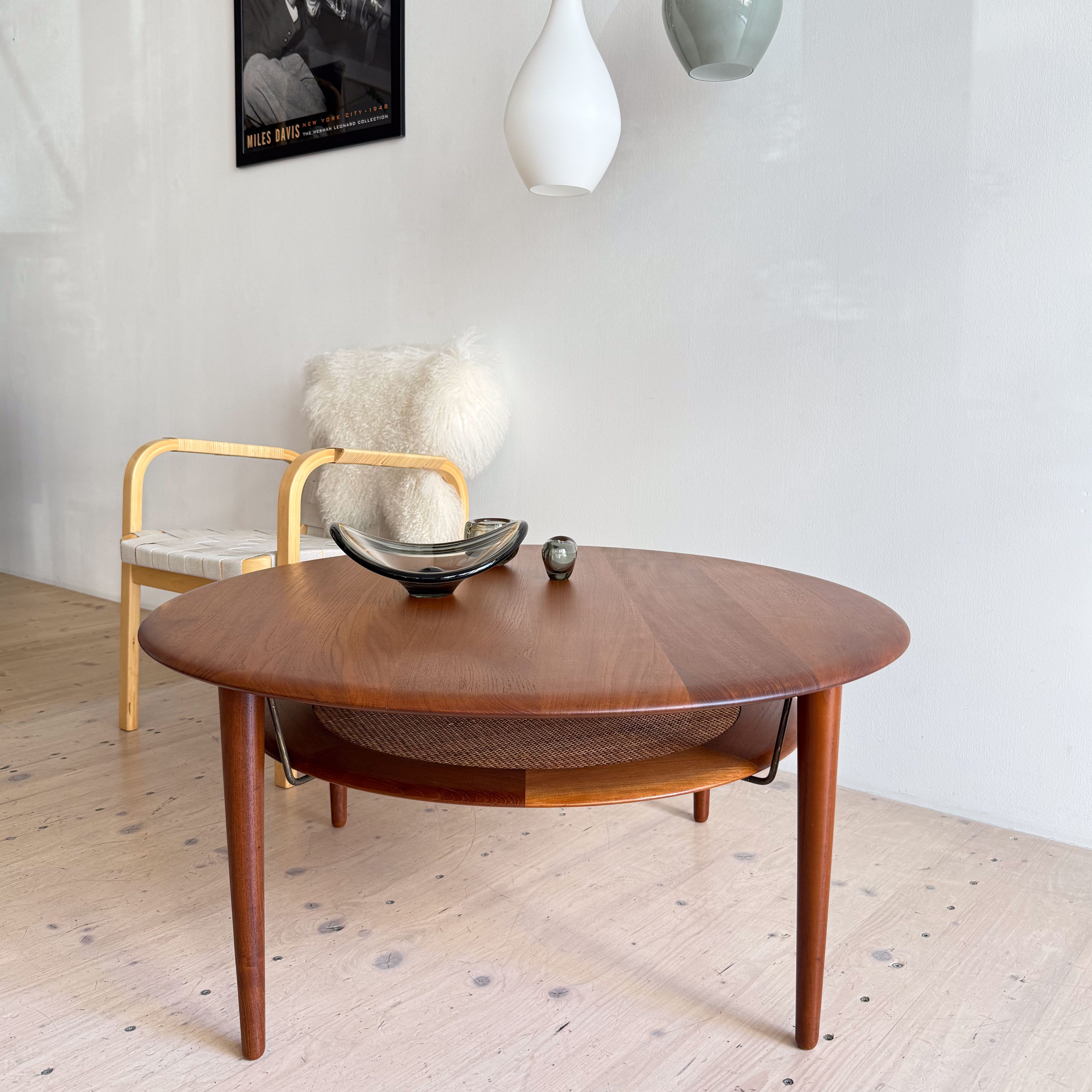 A modern living room scene featuring a round wooden coffee table with a decorative bowl and a small object on top, a light-colored armchair with a fluffy cushion beside it, and abstract wall art in the background.