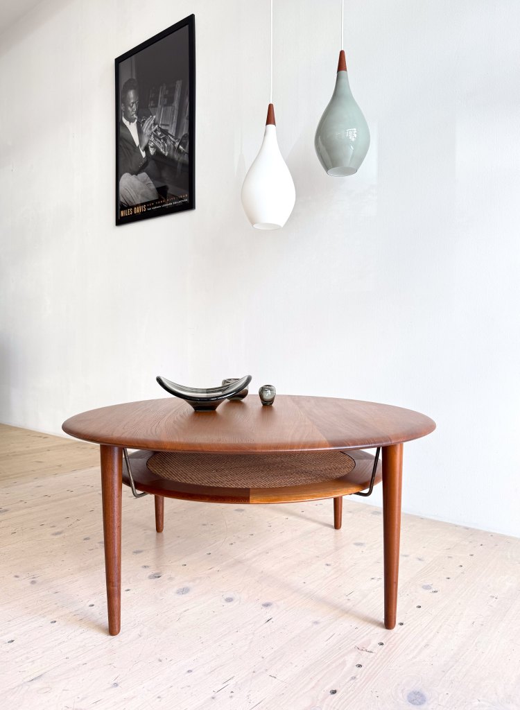 A stylish wooden coffee table with a round top and slatted bottom shelf, accompanied by decorative items on top. Above, two modern pendant lights hang from the ceiling, and a black and white poster of a musician is on the wall.