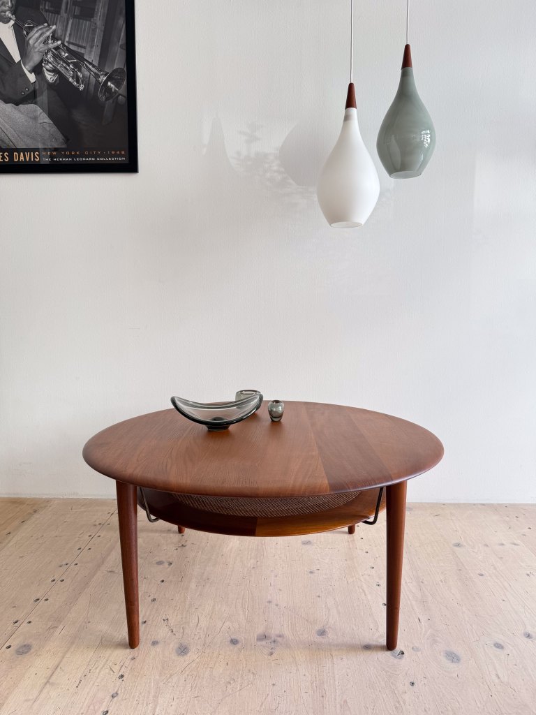 A mid-century modern round wooden coffee table with slender legs, featuring a decorative woven detail on one side, topped with a sleek glass bowl and a small decorative stone. Two uniquely shaped pendant lights hang above against a light-colored wall, with a black and white poster of a musician in the background.