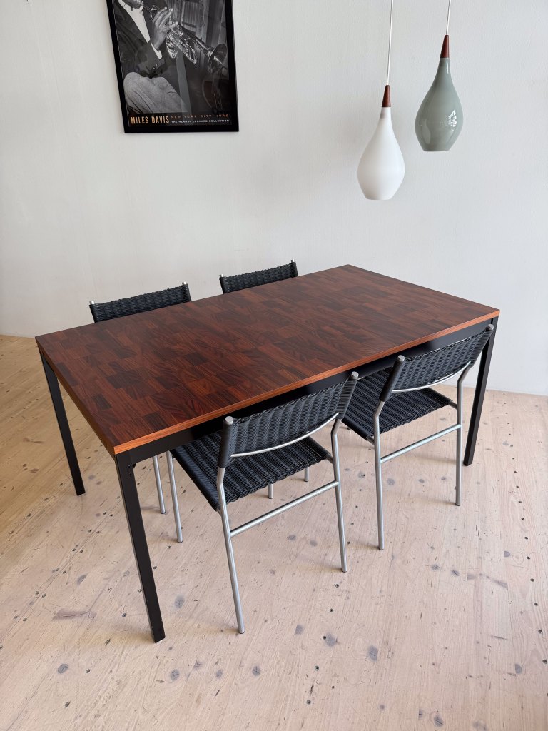 A wooden dining table with a dark patterned surface and four black woven chairs around it, set in a minimalist room with pendant lighting and a jazz music poster on the wall.