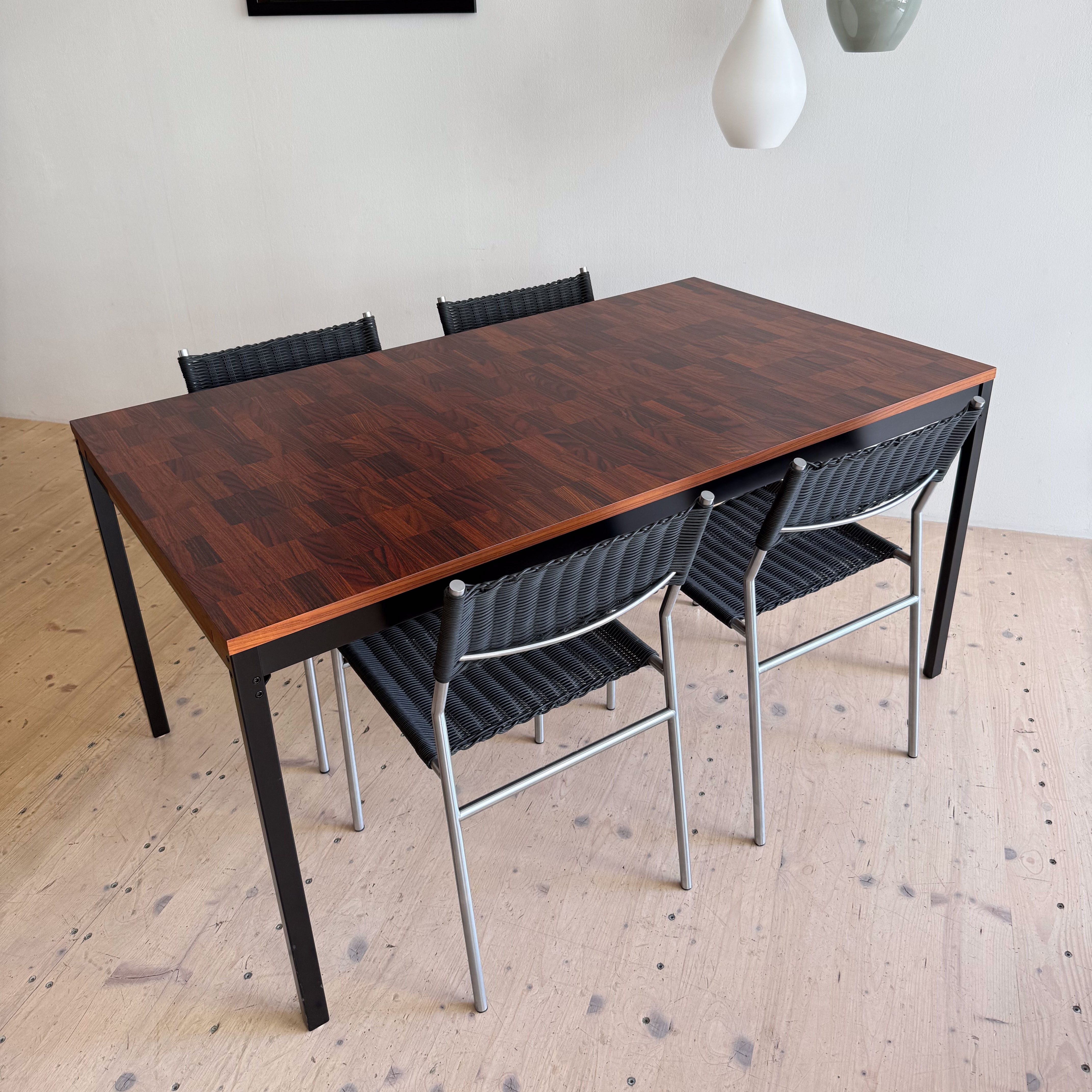 A modern dining table with a dark wooden top and metal legs, surrounded by four black woven chairs. Two pendant lights hang above, and a black and white poster of a musician is visible on the wall.