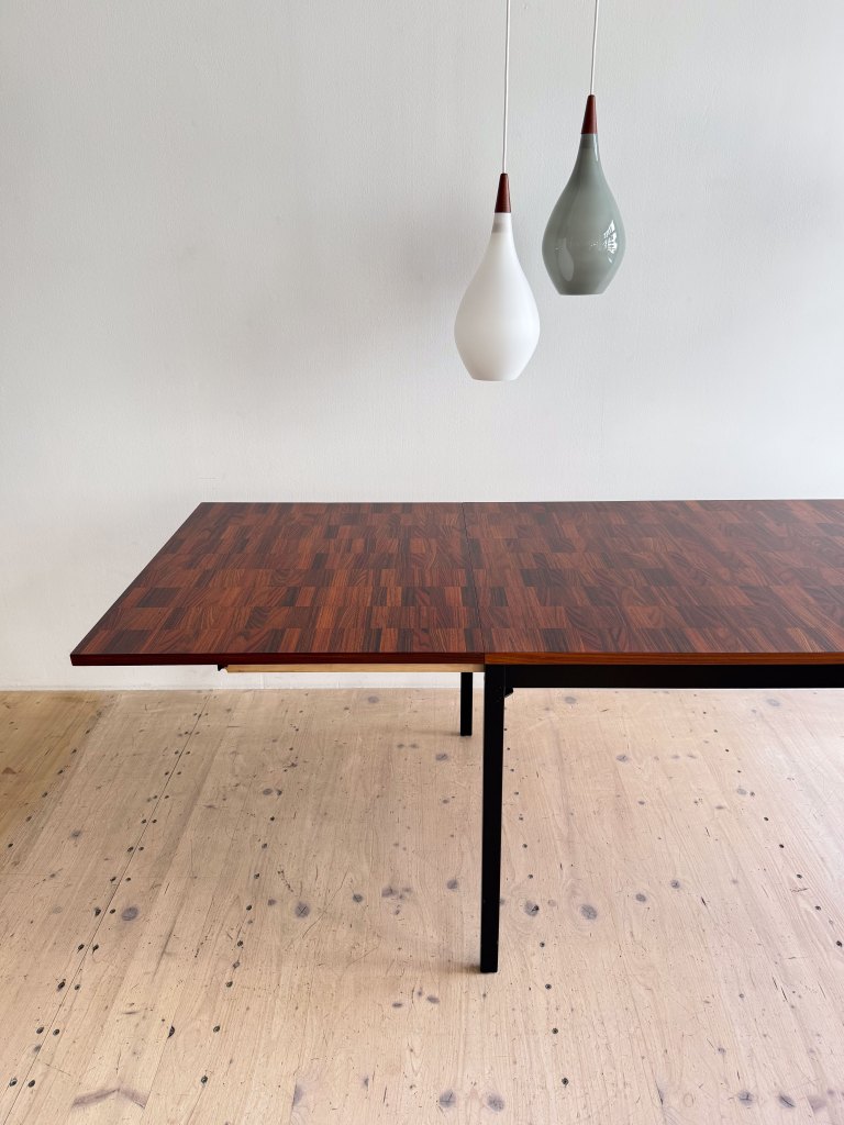 A modern wooden dining table with a patterned surface, accompanied by two hanging pendant lights in white and green above it, set against a neutral wall.