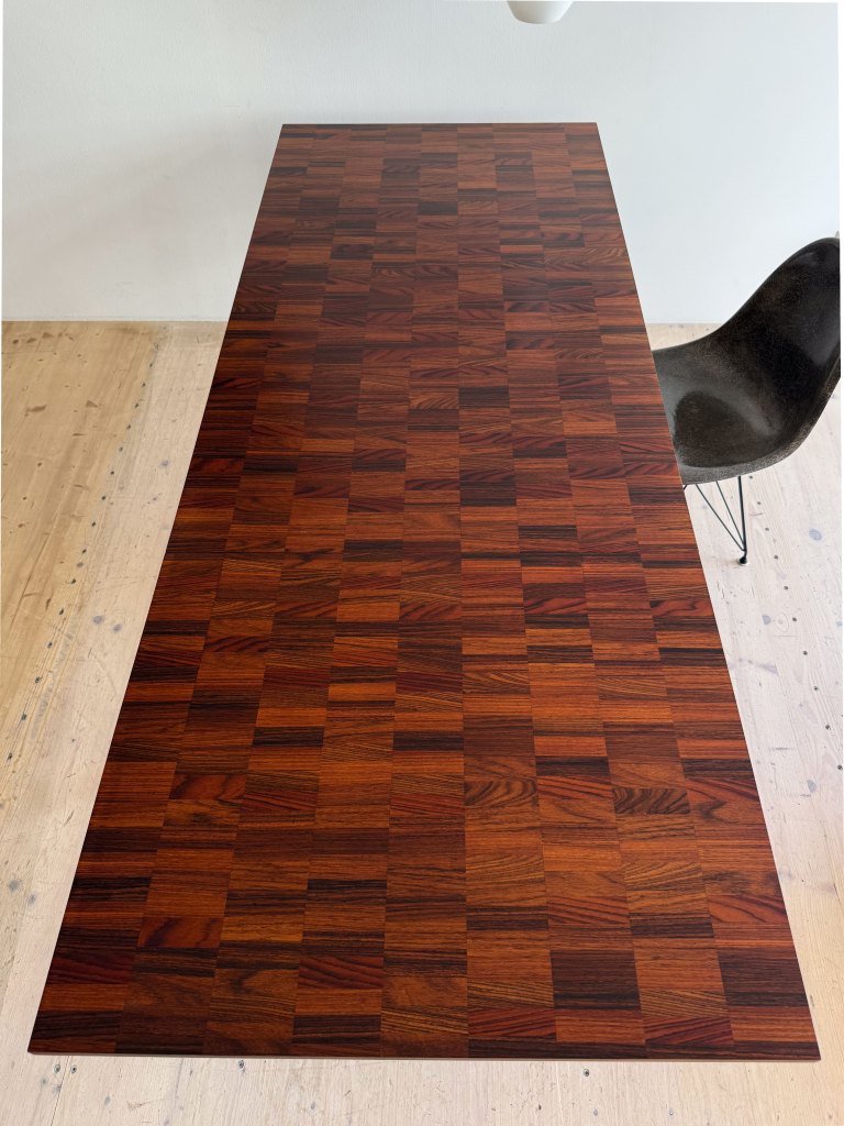A top view of a wooden table with a patchwork design featuring rich, dark brown tones and a smooth finish. A black chair with a unique shape is positioned beside the table, and light-colored wooden flooring is visible beneath.