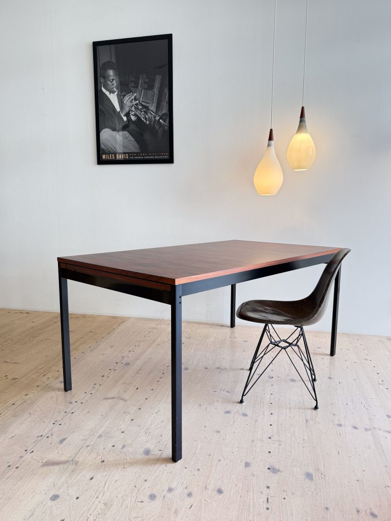 A modern dining table with a wooden top and black legs, a stylish chair beside it, and hanging pendant lights. A black and white framed poster of a musician is mounted on the wall.