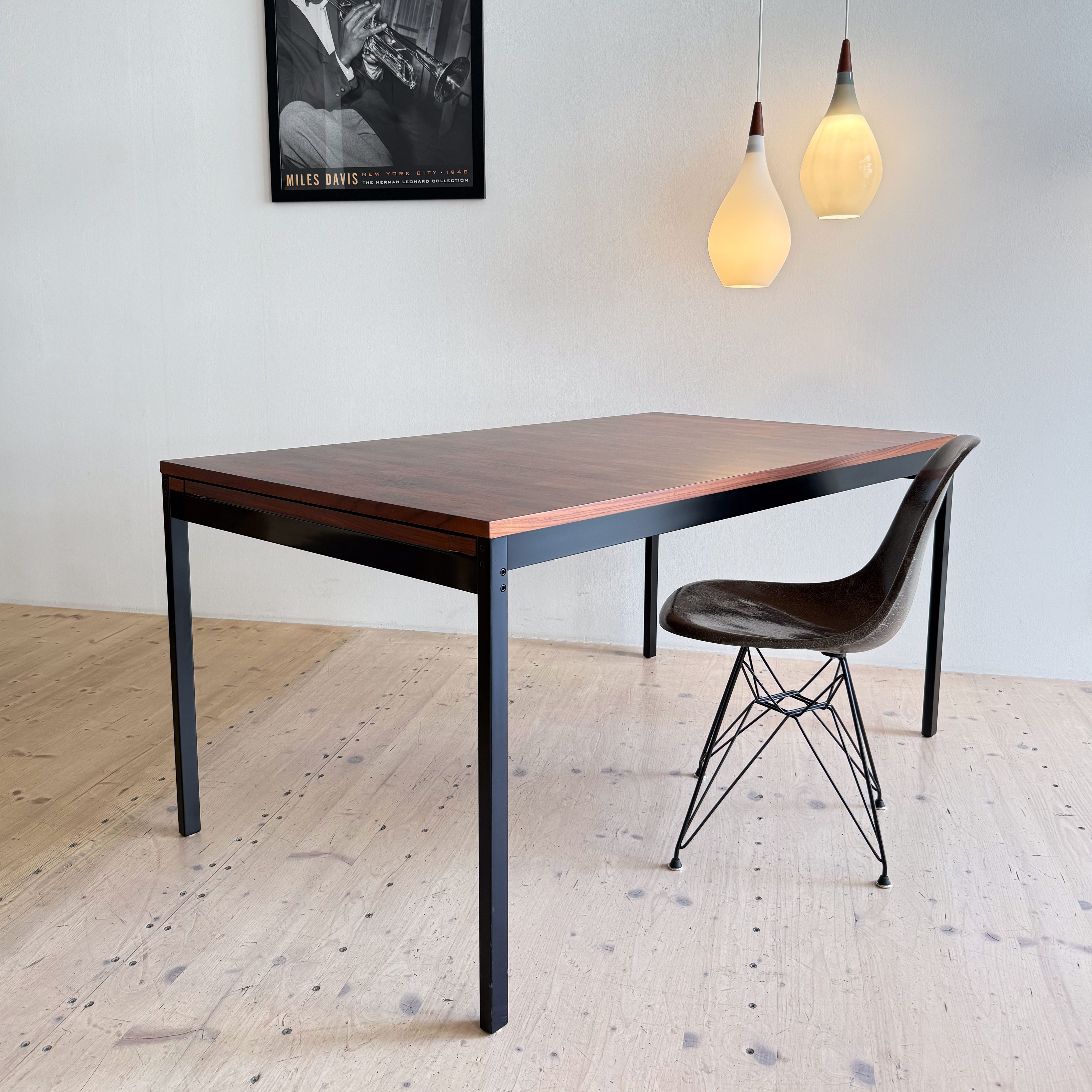 A modern dining table with a wooden surface and sleek black metal legs, accompanied by a stylish chair with a wire base. Above the table, two contemporary light fixtures hang, and a framed black-and-white poster of a musician is displayed on the wall.