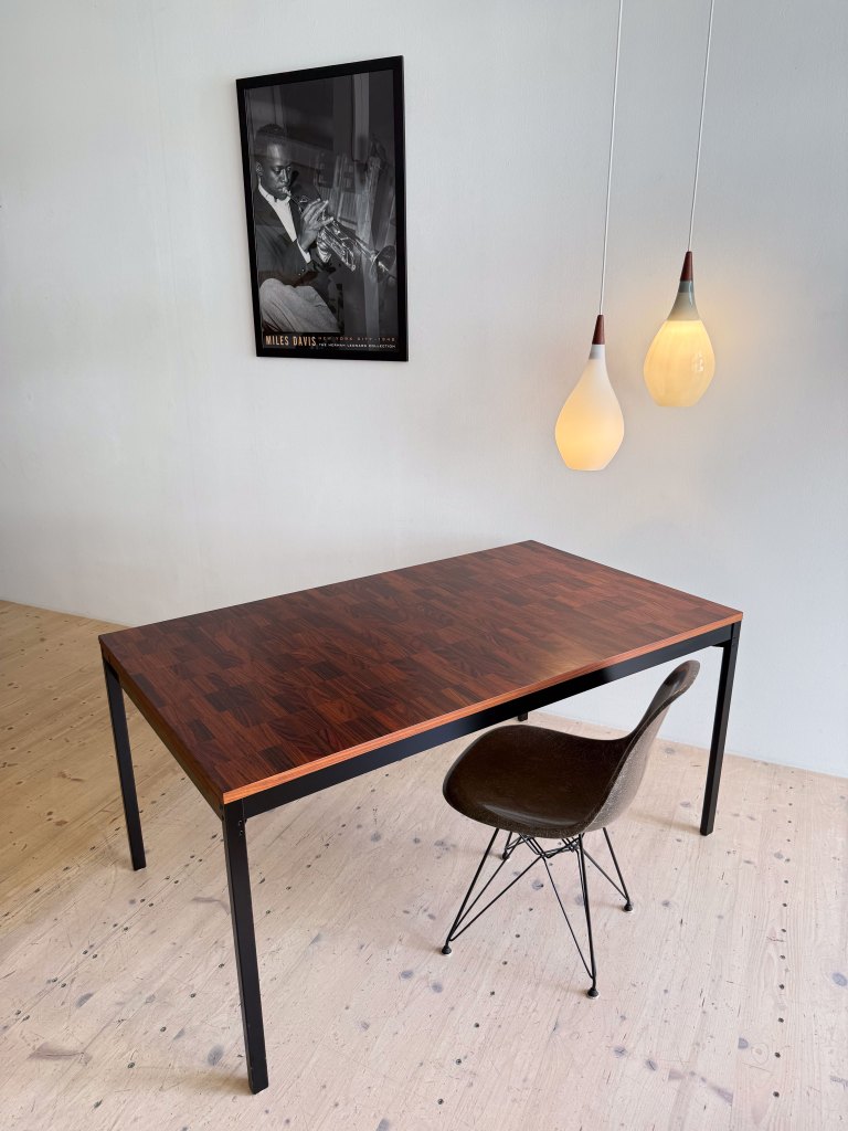 A modern wooden table with a polished surface and an Eames-style chair, beneath two pendant lights, with a black and white poster of a jazz musician on the wall.