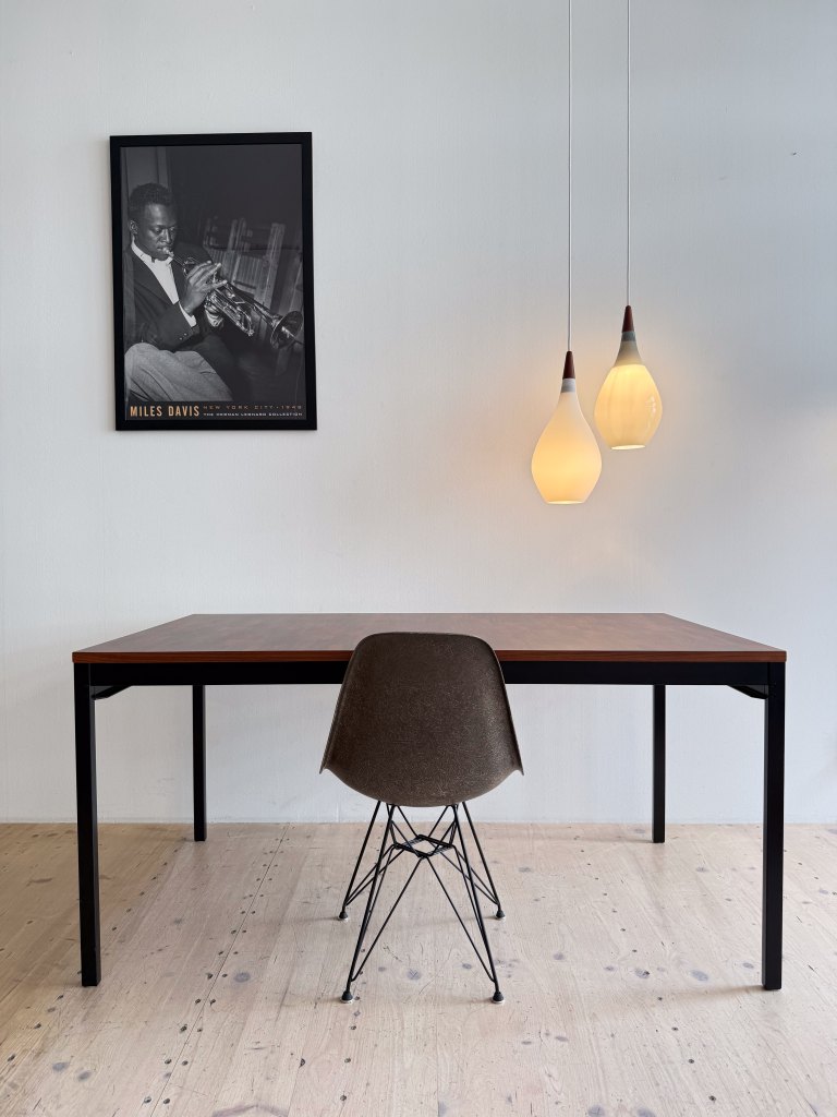 A modern, minimalist workspace featuring a wooden table and a stylish chair. A framed black and white photo of a musician playing the trumpet hangs on the wall, accompanied by three pendant lights that provide warm illumination.