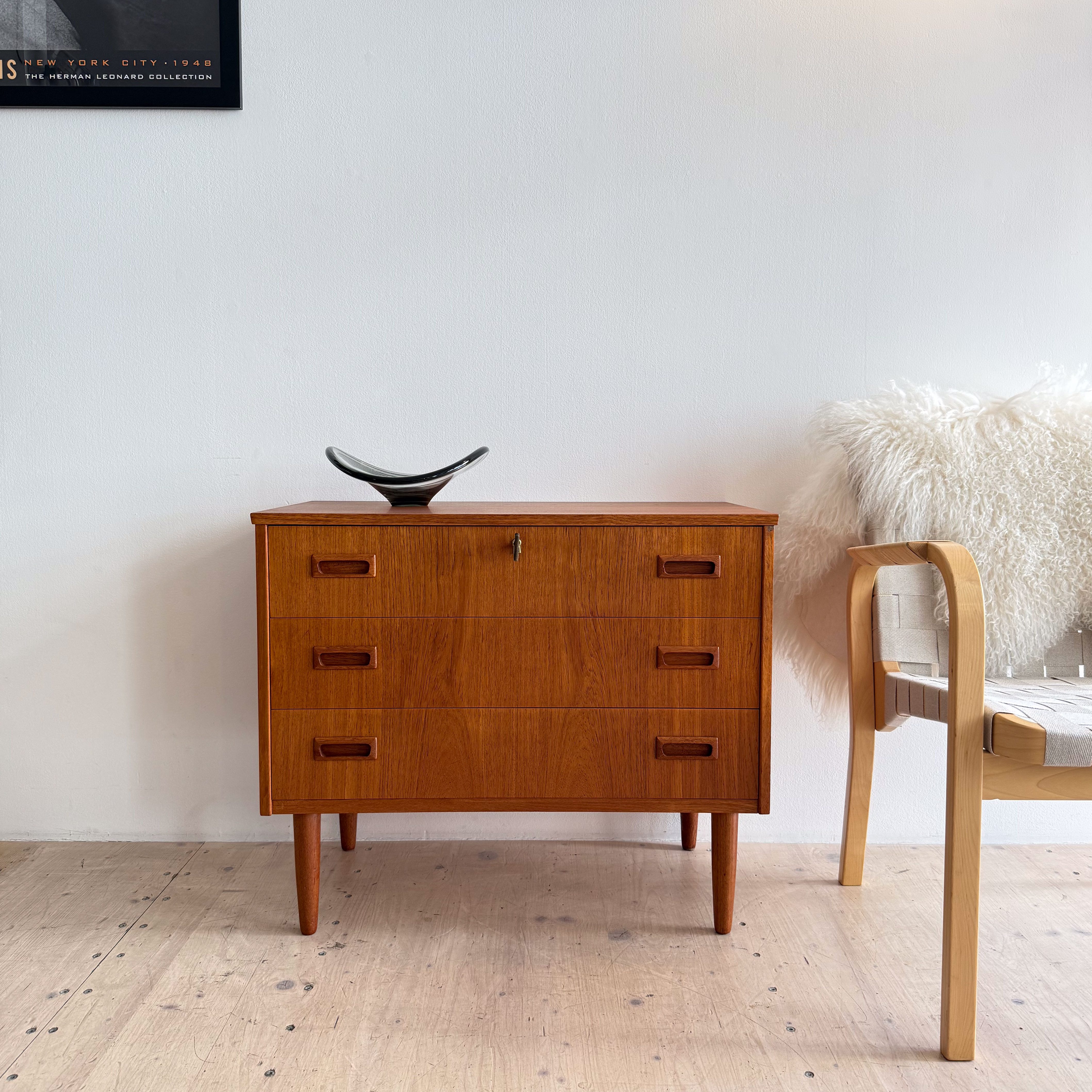 A wooden cabinet with three drawers, featuring a sleek design and wooden handles, placed against a minimalist wall; a decorative bowl sits on top, beside a light-colored chair with a woven seat and a plush throw.