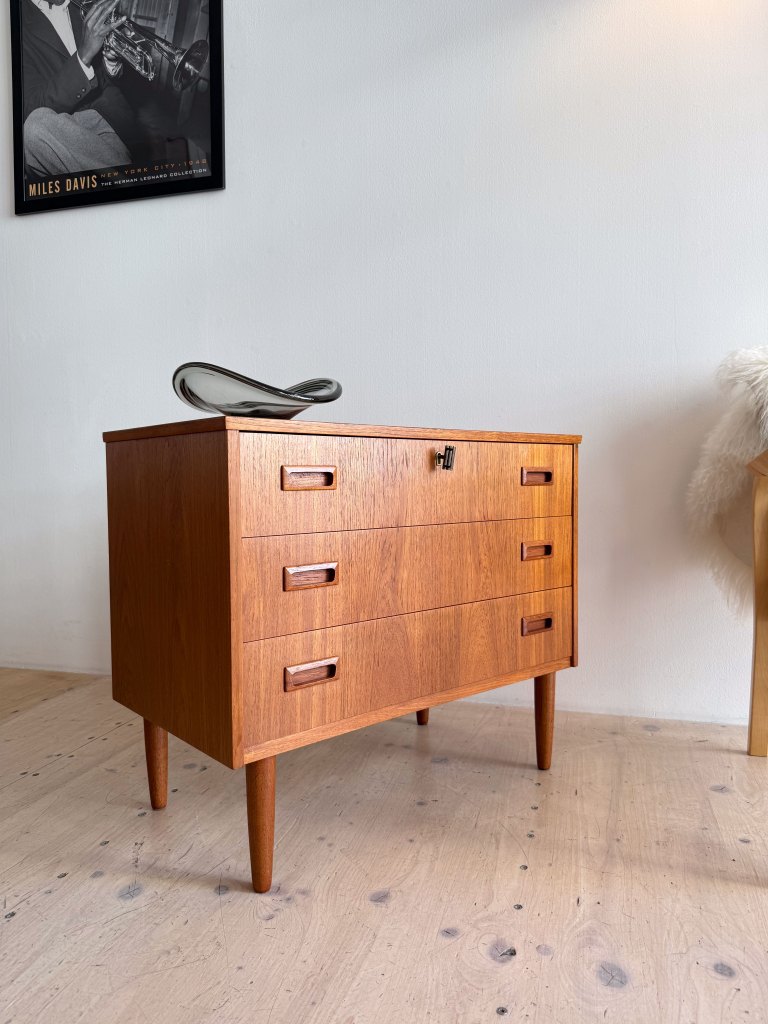 A mid-century modern wooden chest of drawers with four drawers, featuring slender legs and simple metal handles, displayed in a stylish interior.