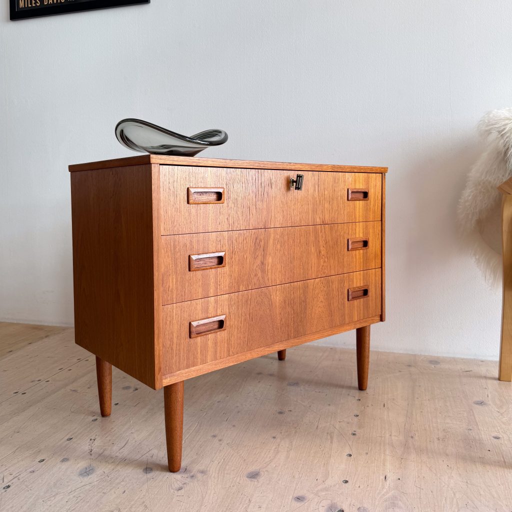 A vintage wooden dresser with three drawers and tapered legs, topped with a decorative glass piece on a neutral wall background.