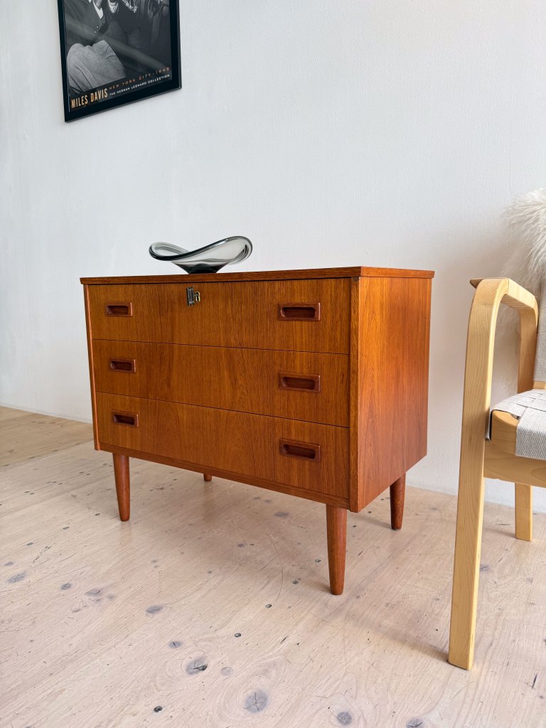A mid-century modern wooden dresser with three drawers and tapered legs, placed against a white wall. A decorative bowl sits on top and a chair is nearby.