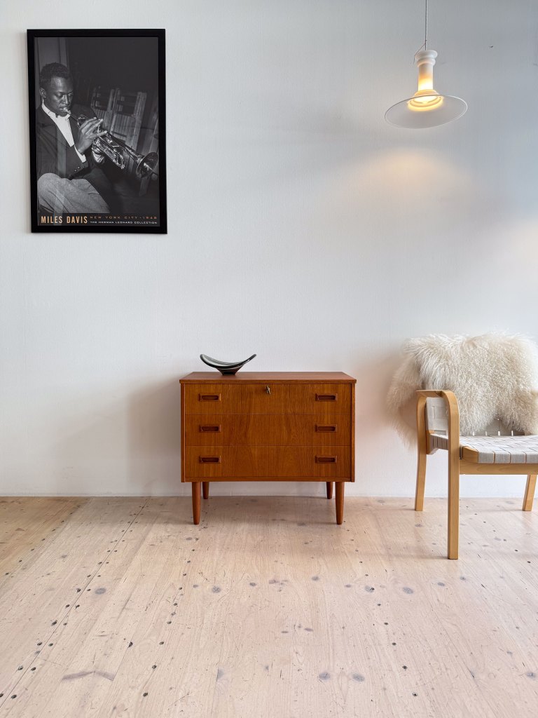 A stylish interior showcasing a wooden chest of drawers, a contemporary chair, and a fur throw. A pendant light hangs above, and a framed black and white photograph of a musician is on the wall.