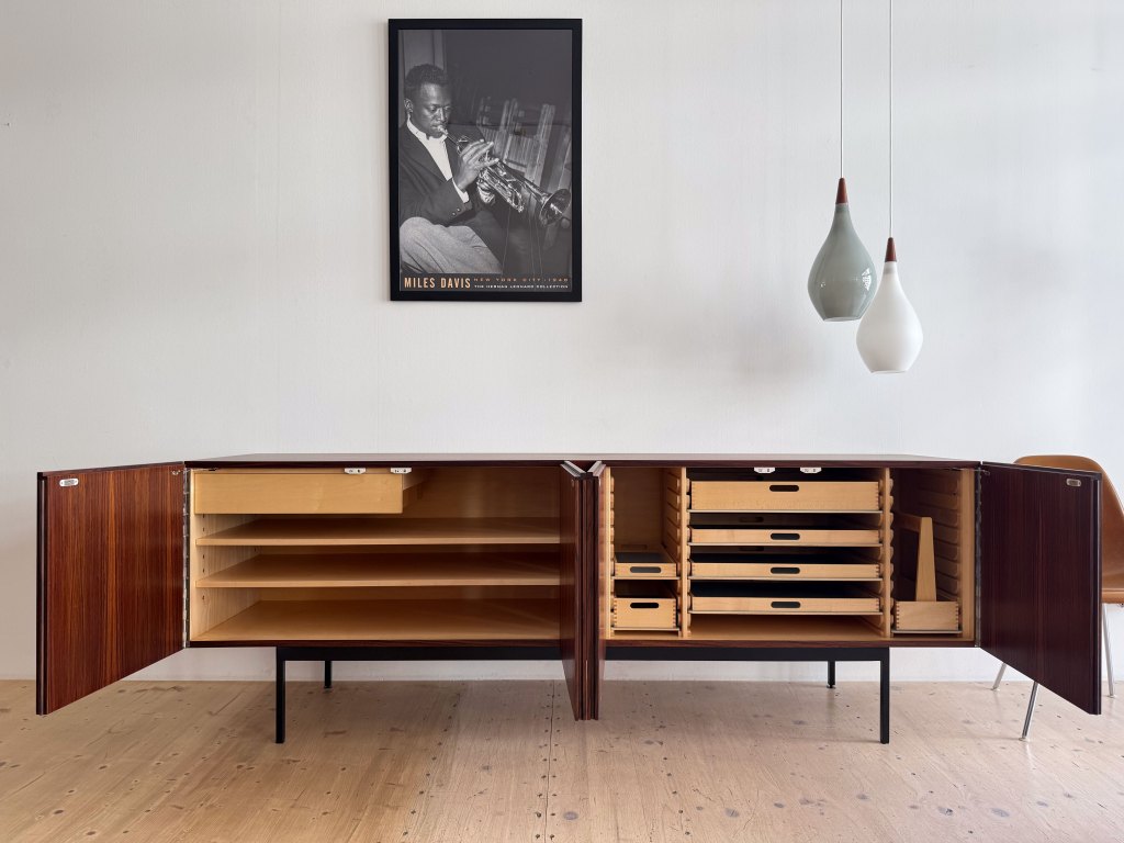 A modern wooden sideboard with open doors revealing shelves and drawers, positioned in a minimalist room with light wood flooring. Above the sideboard hangs a black and white poster of a musician playing the trumpet, featuring the name 'Miles Davis.' Two pendant lights in varying shapes and colors are suspended from the ceiling.