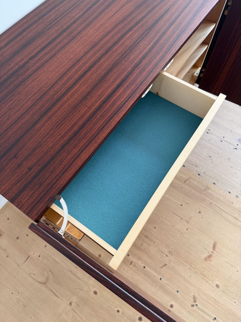 A close-up view of a wooden desk with a rich, dark wood finish. An open drawer reveals a light-colored interior with a teal lining.