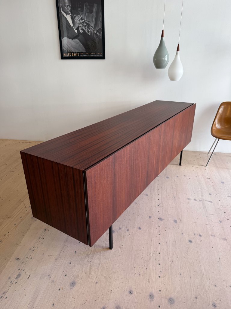 A modern wooden sideboard with a sleek design, featuring a rich brown finish and minimalist black legs, positioned on a light wooden floor. Decorative pendant lights hang above, and a framed poster of a musician is visible on the wall behind.