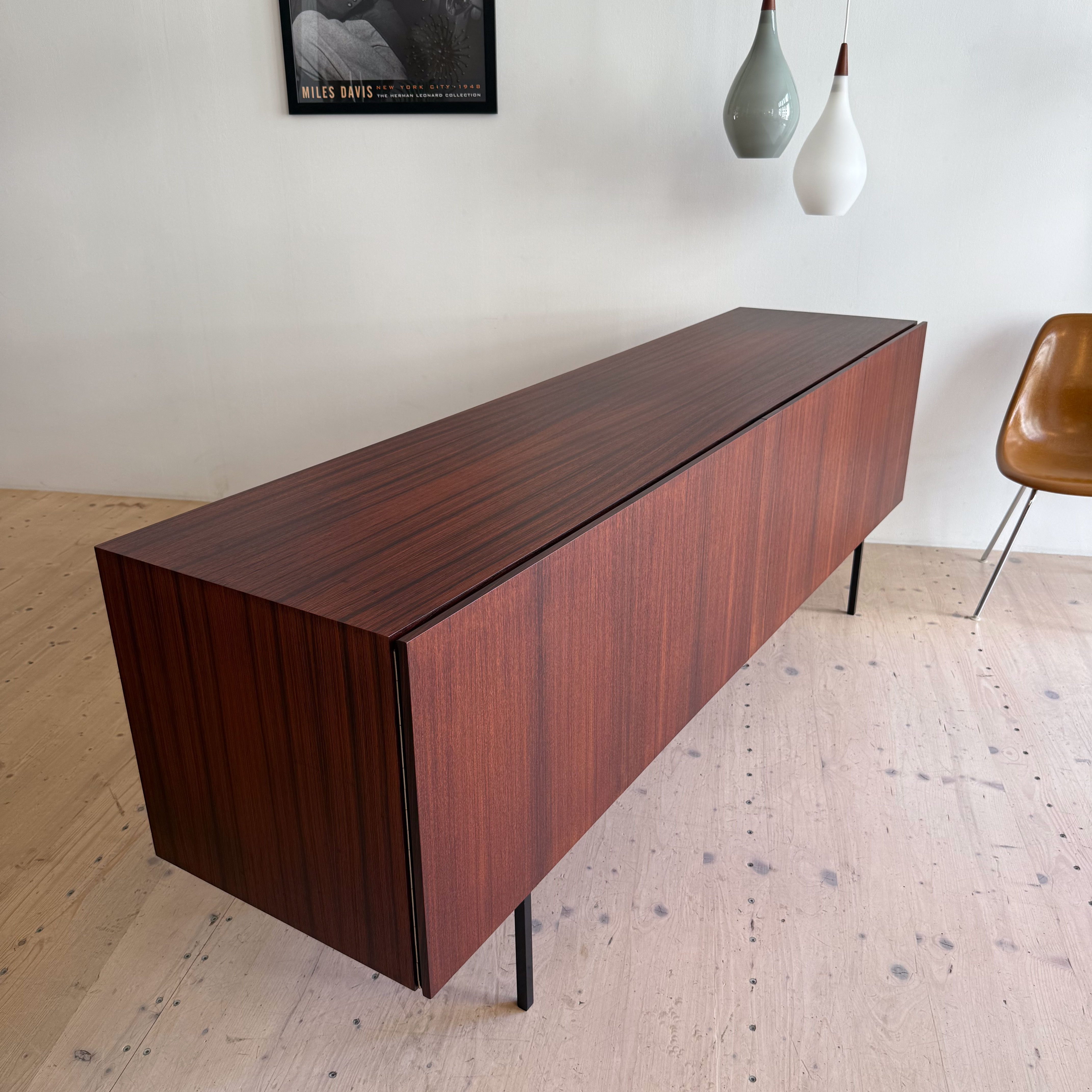 A modern wooden sideboard in a rich brown finish, featuring clean lines and standing on thin black legs, with two suspended light fixtures and a vintage jazz poster in the background.