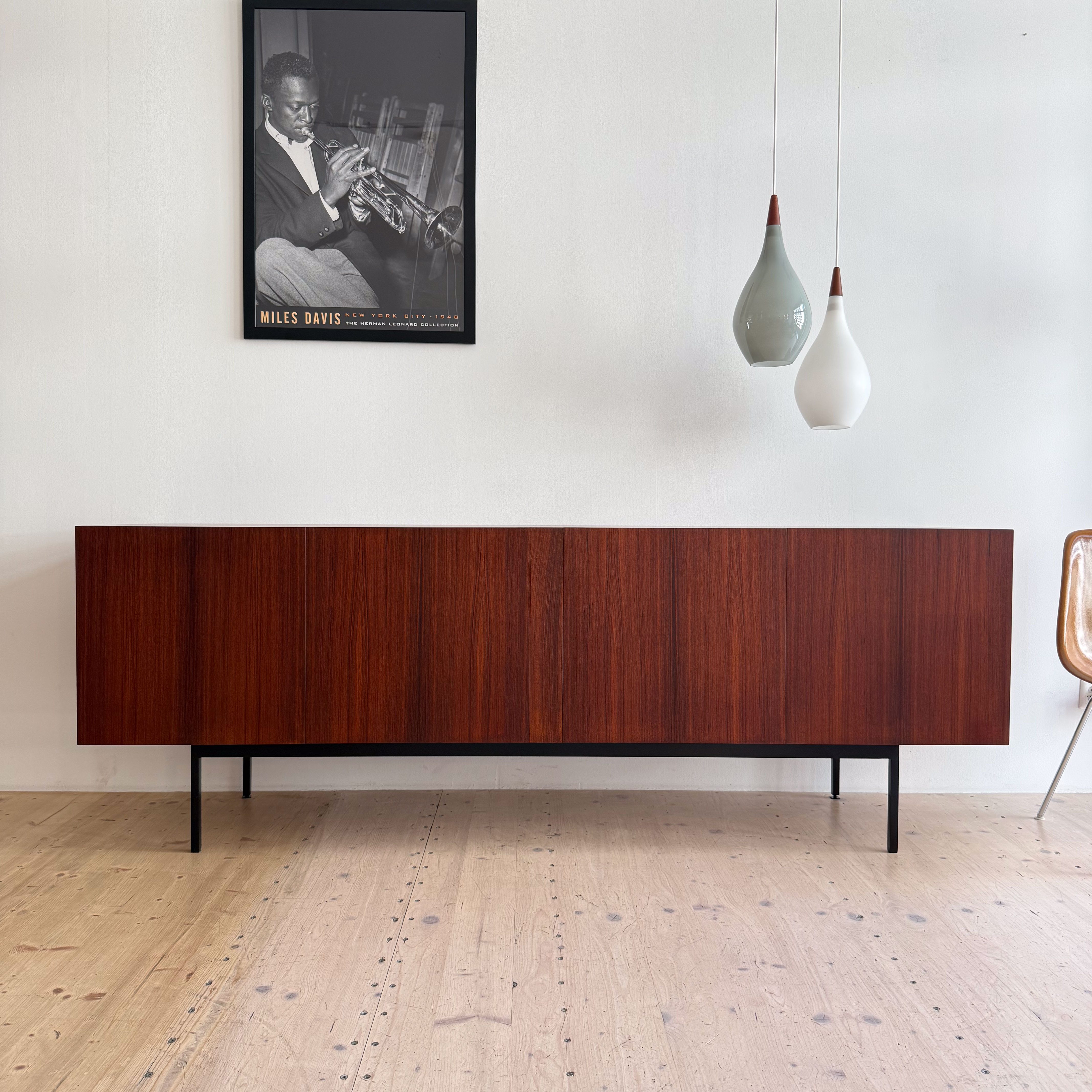 A modern, low wooden cabinet with a rich brown finish, sitting on slender black legs, against a white wall. Above it hangs a framed black and white poster of a musician playing the trumpet, with decorative hanging light fixtures in varying shapes and colors.