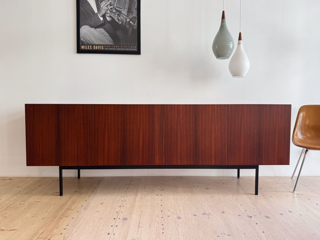 A modern wooden sideboard with a rich, dark finish, featuring sliding doors and sleek black legs, positioned against a white wall. Above it hangs a framed poster of Miles Davis, and there are two minimalist pendant lights in soft colors. A mid-century modern chair is partially visible to the right.