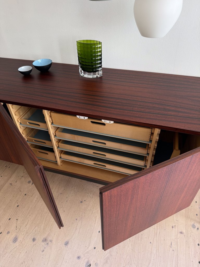 A wooden sideboard with open doors revealing multiple drawers, topped with a green glass vase and two small bowls.