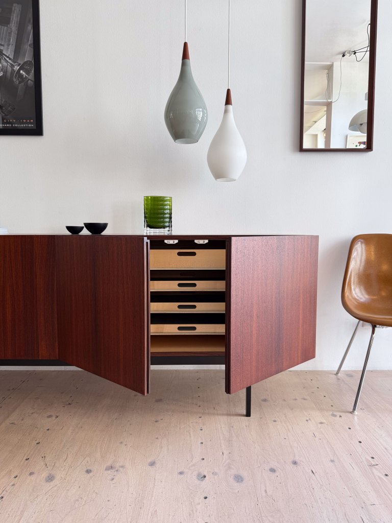 A modern wooden cabinet with open drawers, accompanied by pendant lights in various colors. A green glass vase and small black bowls are placed on top, next to a mid-century style chair and a mirror reflecting the room.