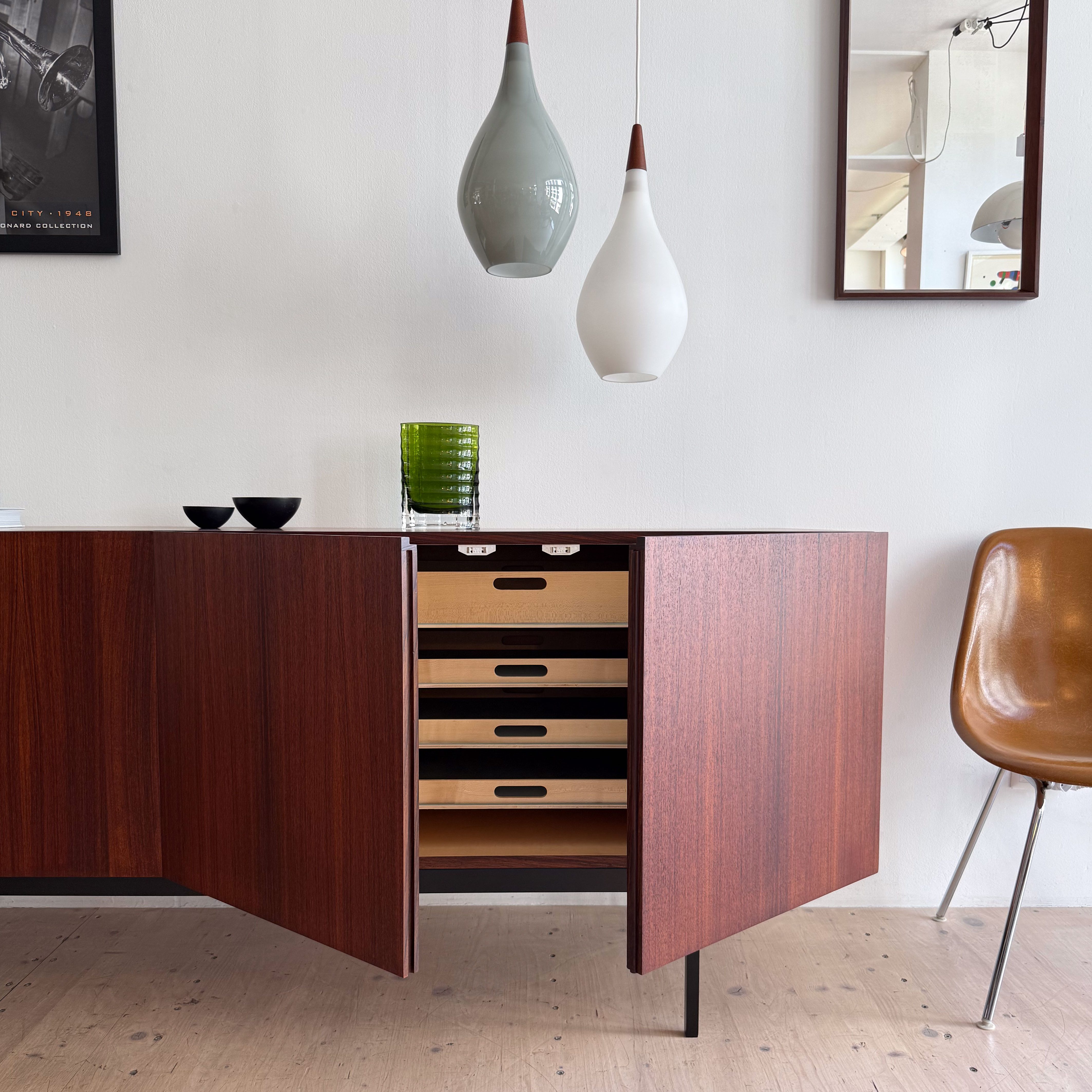 A wooden sideboard with open doors revealing drawers, accompanied by two hanging pendant lights and a green glass vase on top, set against a light-colored wall.