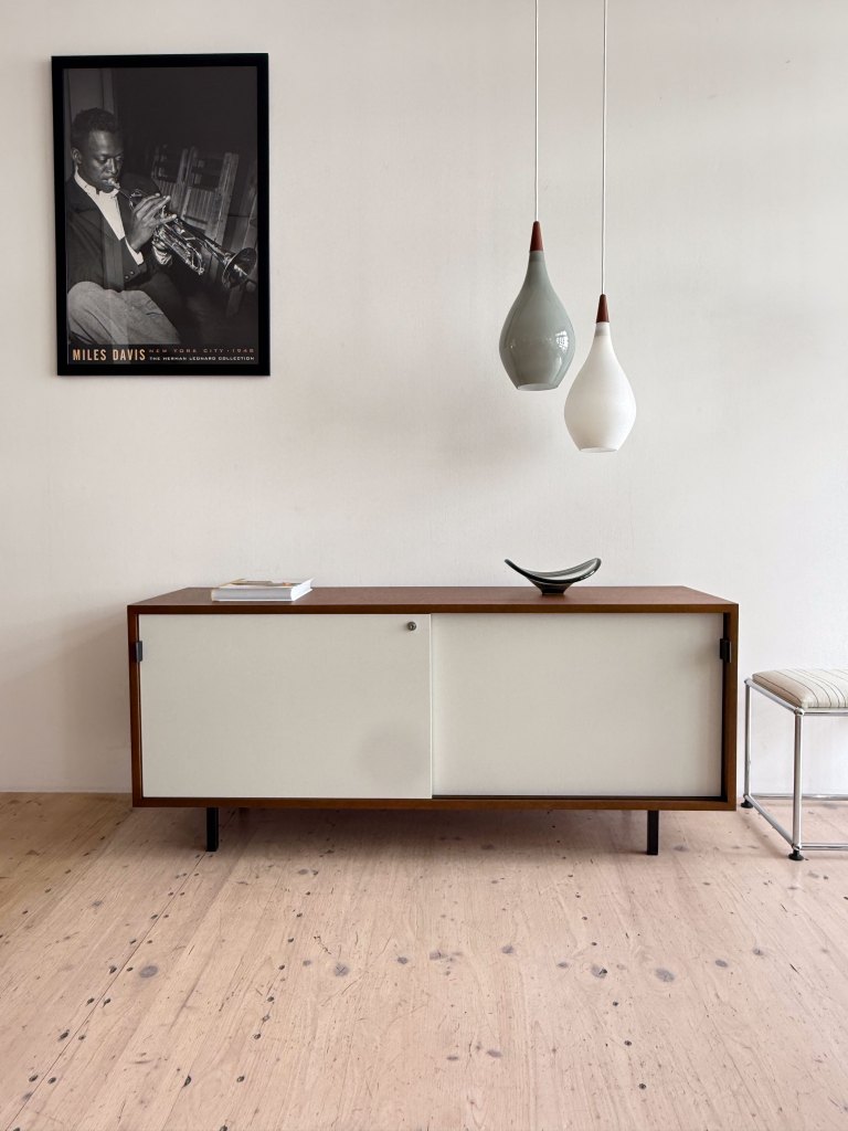 A modern wooden sideboard with white sliding doors, accompanied by a decorative bowl. Above, three hanging pendant lights in varying shapes, and a black-and-white framed poster of a trumpet player on the wall.