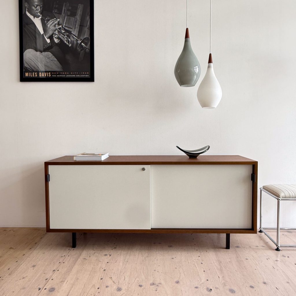 A modern wooden sideboard with a white sliding door, featuring a small decorative dish on top and a book. Two pendant lights hang above in different colors, with a poster of a jazz musician on the wall behind.