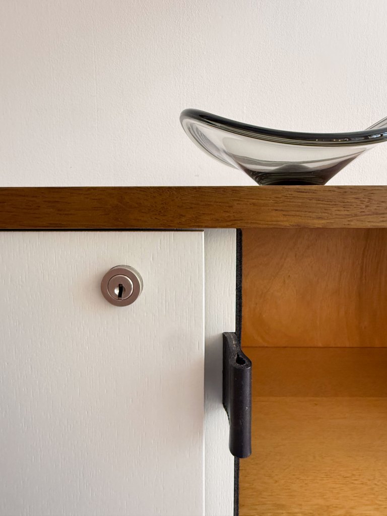 Close-up of a wooden cabinet with a white door featuring a keyhole and a black handle, with a glass dish resting on top.