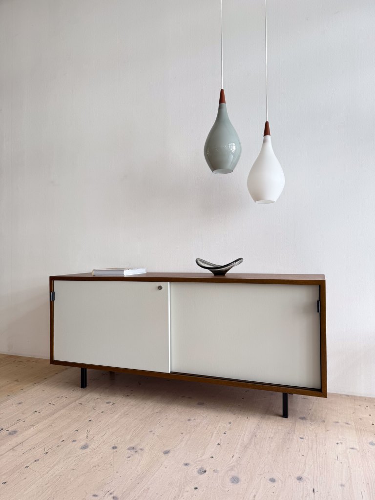 A modern wooden sideboard with a sliding door, featuring a white front and black legs, accompanied by a decorative dish on top and two hanging light fixtures overhead.