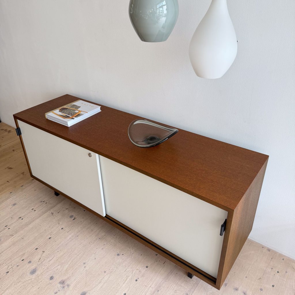 A modern wooden cabinet with two sliding white doors, a magazine on top, and a decorative glass dish, set against a light wall.