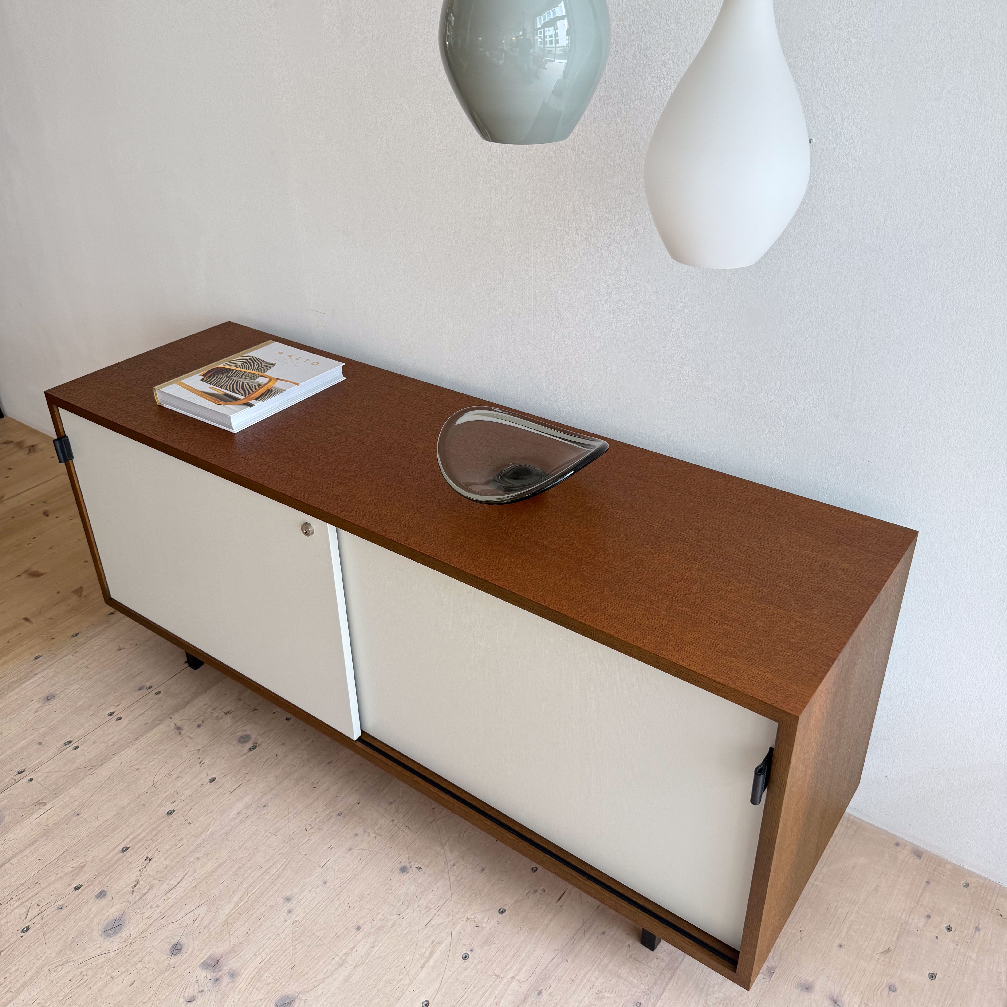 A modern wooden sideboard with a combination of dark wood and white sliding doors, topped with a decorative glass vase and magazines, under two pendant light fixtures.