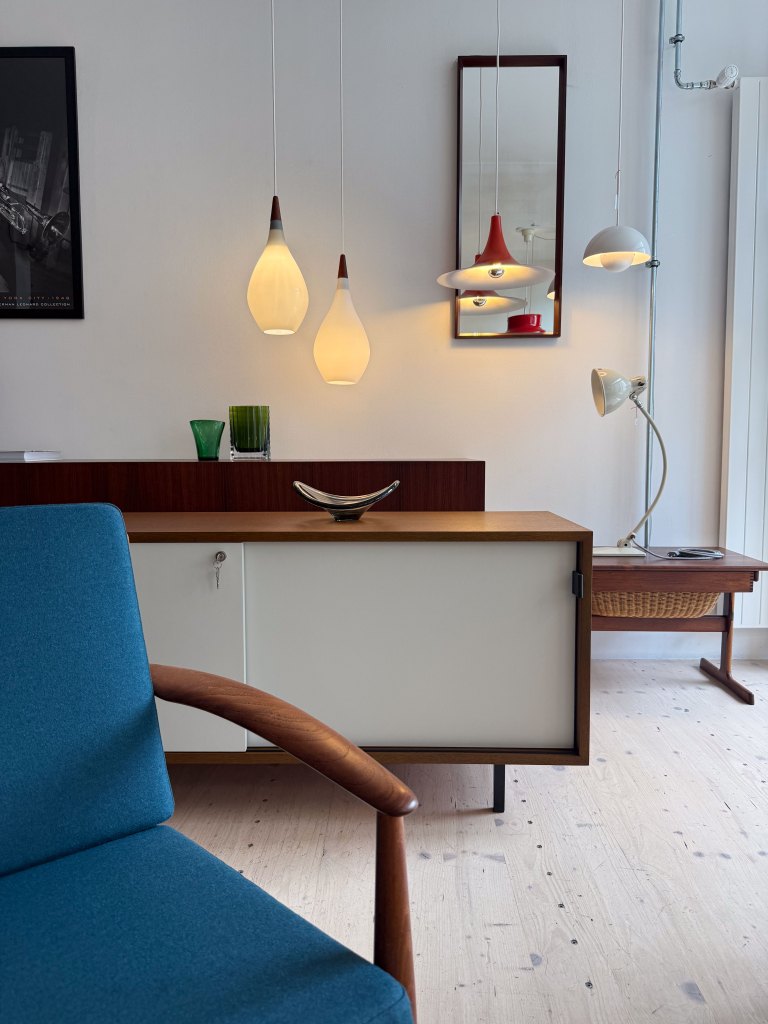 A modern living space featuring a blue upholstered chair, a wooden sideboard with white cabinet doors, decorative glass vases, and various pendant lights hanging from the ceiling. A mirror is mounted on the wall, reflecting the elegant decor.