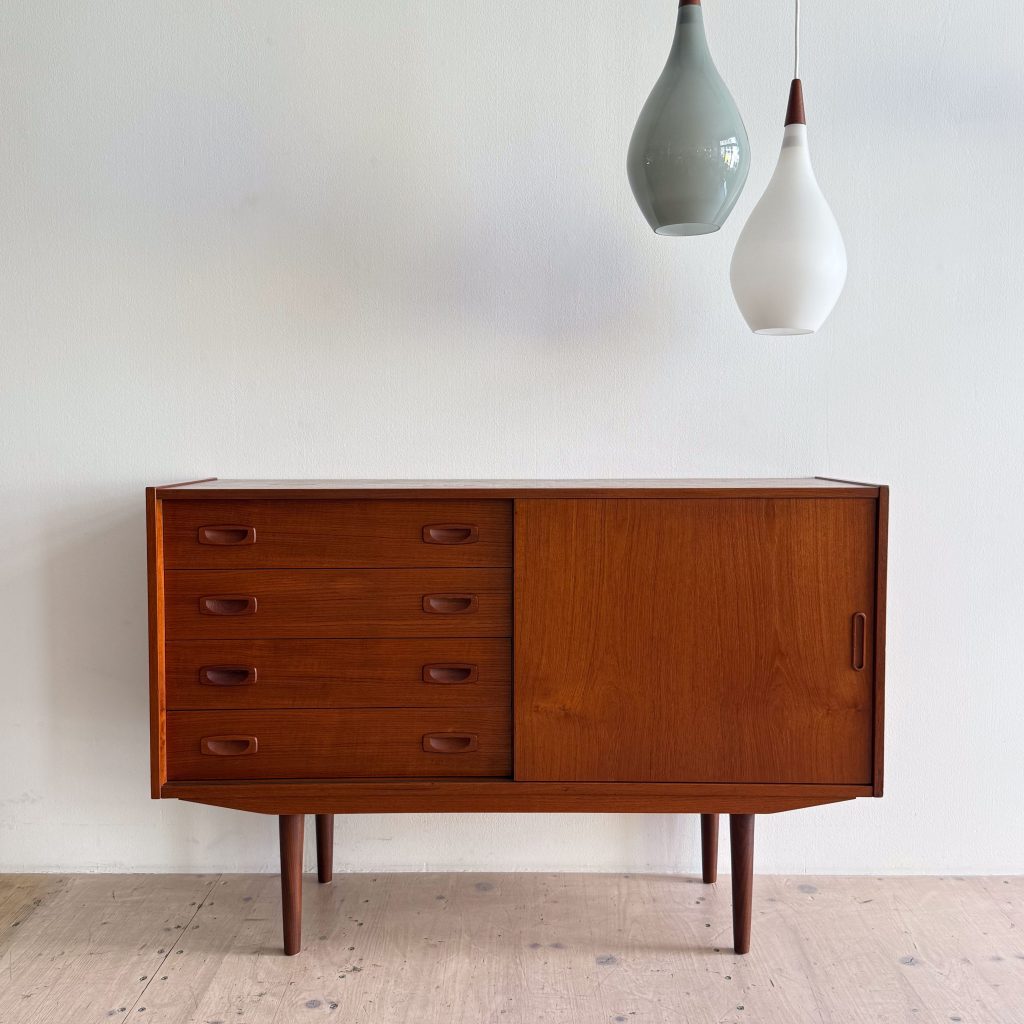 A mid-century modern wooden sideboard with four drawers and a cabinet door, featuring sleek lines and splayed legs, accompanied by two hanging pendant lights.