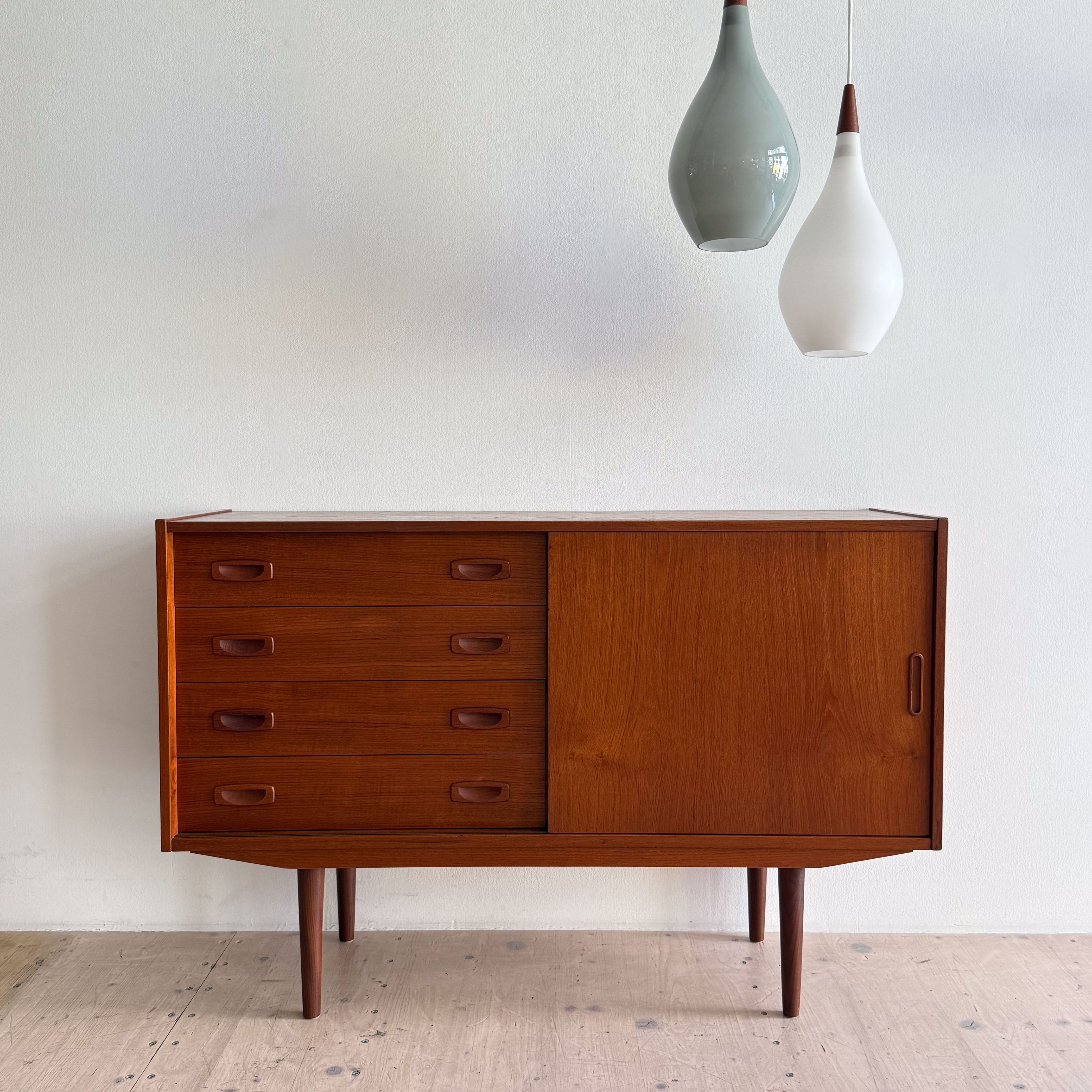 Mid-century modern wooden cabinet with six drawers and sliding door, accompanied by two pendant lights in different colors.