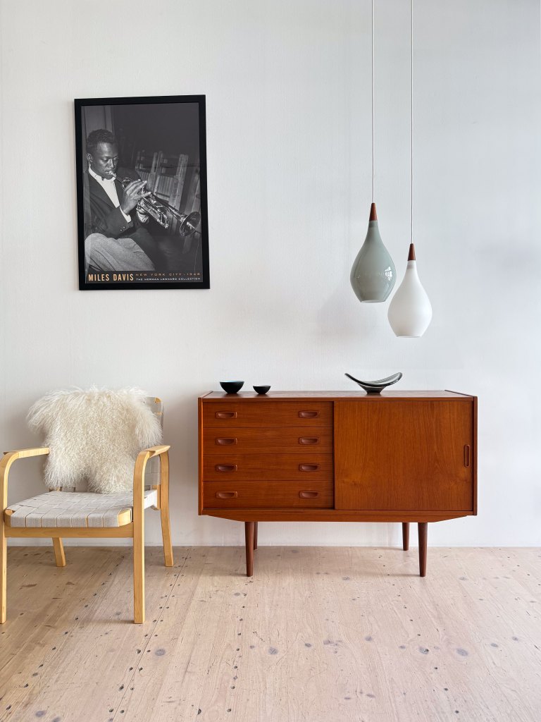 A mid-century modern wooden sideboard with multiple drawers and a sliding door, topped with decorative bowls. Beside it is a light wooden chair with a plush white fur throw. Above the sideboard hangs a black-and-white photo of a musician playing a trumpet.