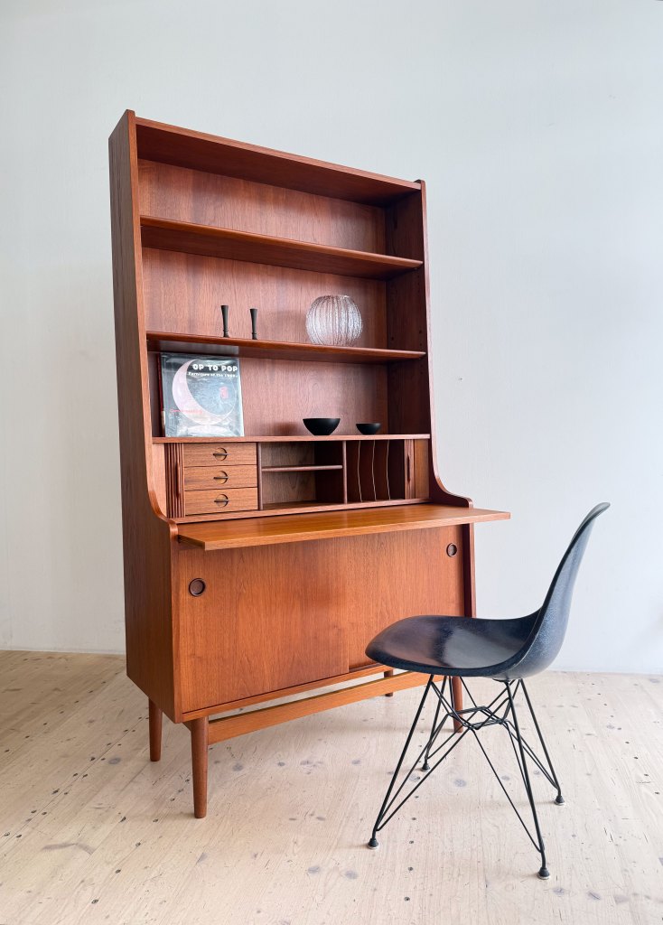 A mid-century modern wooden bookshelf with an attached desk, featuring open shelves displaying decorative items, and a sleek black chair in front.