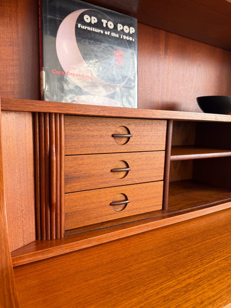 A close-up of a mid-century modern wooden furniture piece featuring three small drawers with circular handles, a book titled 'Op to Pop: Furniture of the 1960s' on the back shelf, and a small black bowl.