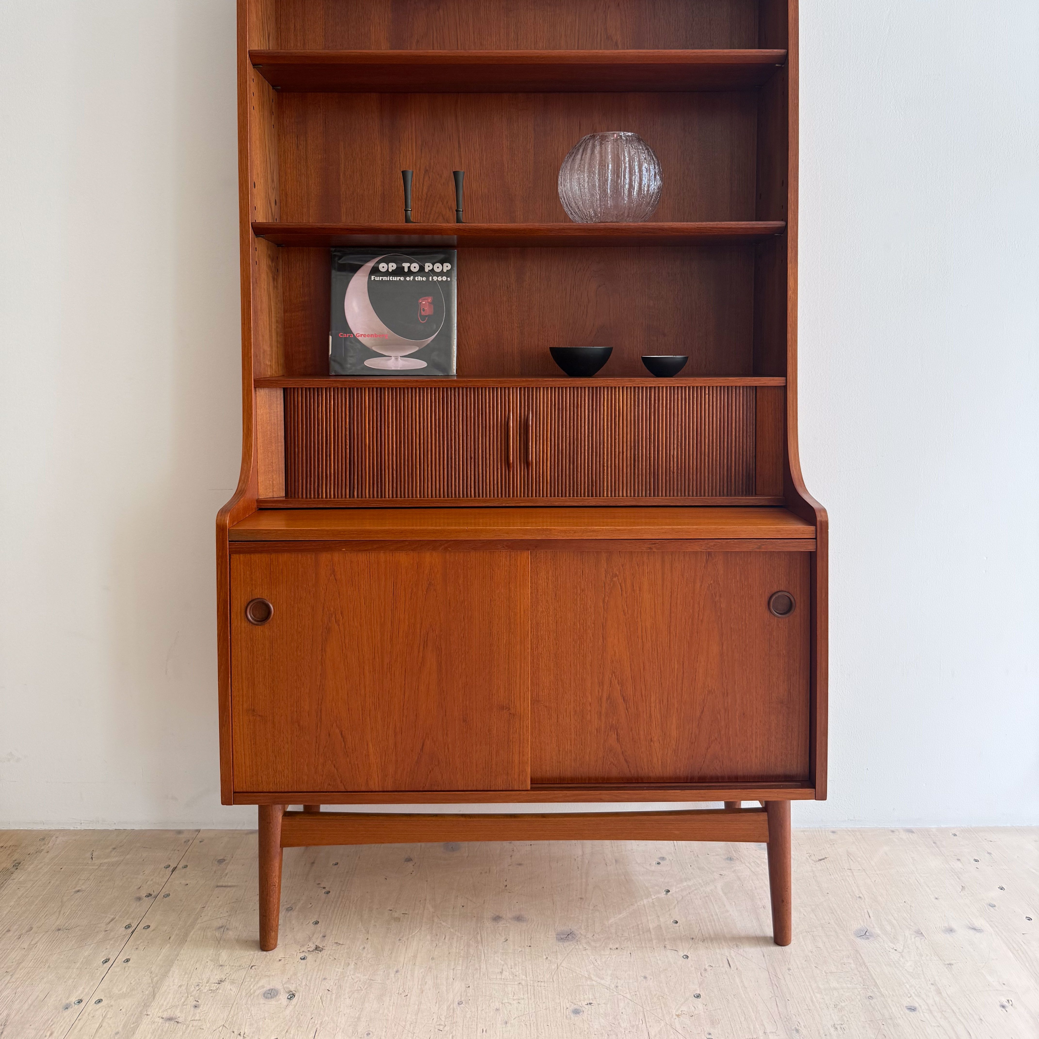 A wooden mid-century modern bookshelf with shelves displaying decorative items, including a round vase and two small bowls, alongside a book. The bottom section features sliding doors and is supported by four legs.