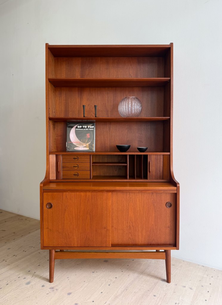 A wooden mid-century modern cabinet with open shelves, featuring decorative items like a vase and bowls, alongside a book on display. The cabinet has a lower sliding door compartment and small drawers.