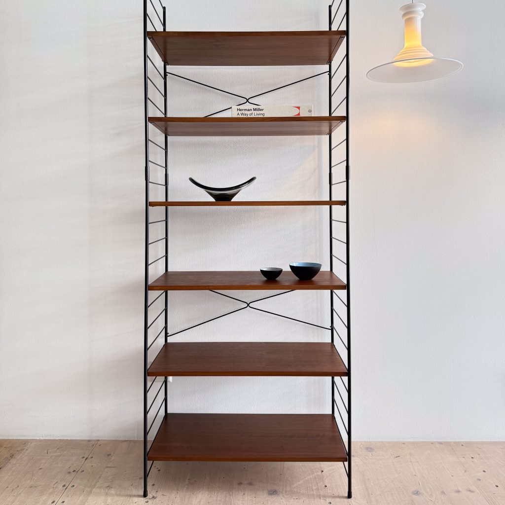 A modern wooden bookshelf with metal frames, featuring several shelves. The middle shelves display a black ceramic bowl and a book titled 'Herman Miller: A Way of Living.' A white pendant light is visible in the upper right corner.