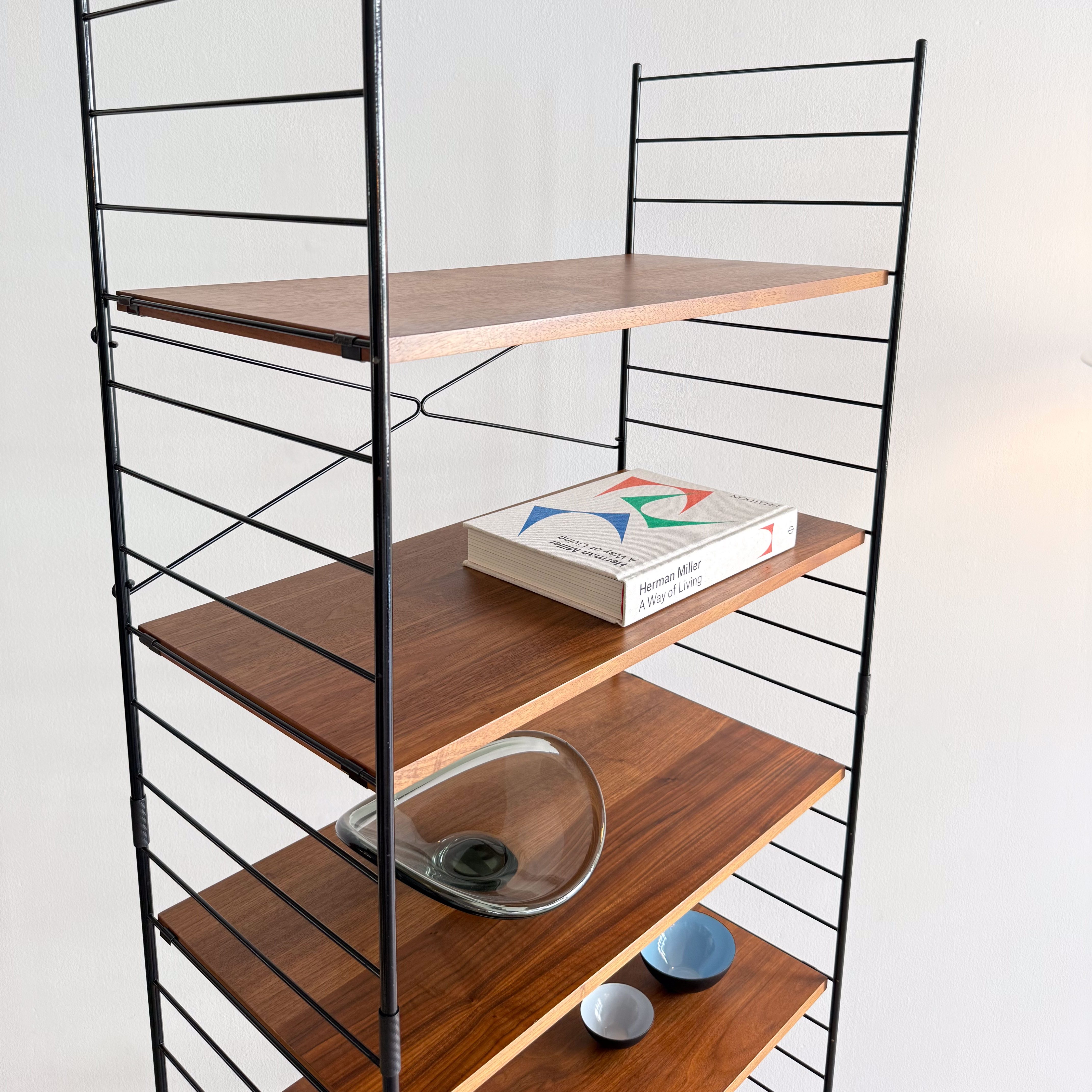 A minimalist wooden shelving unit with black metal frame, displaying a book, a glass bowl, and several small plates on its shelves.
