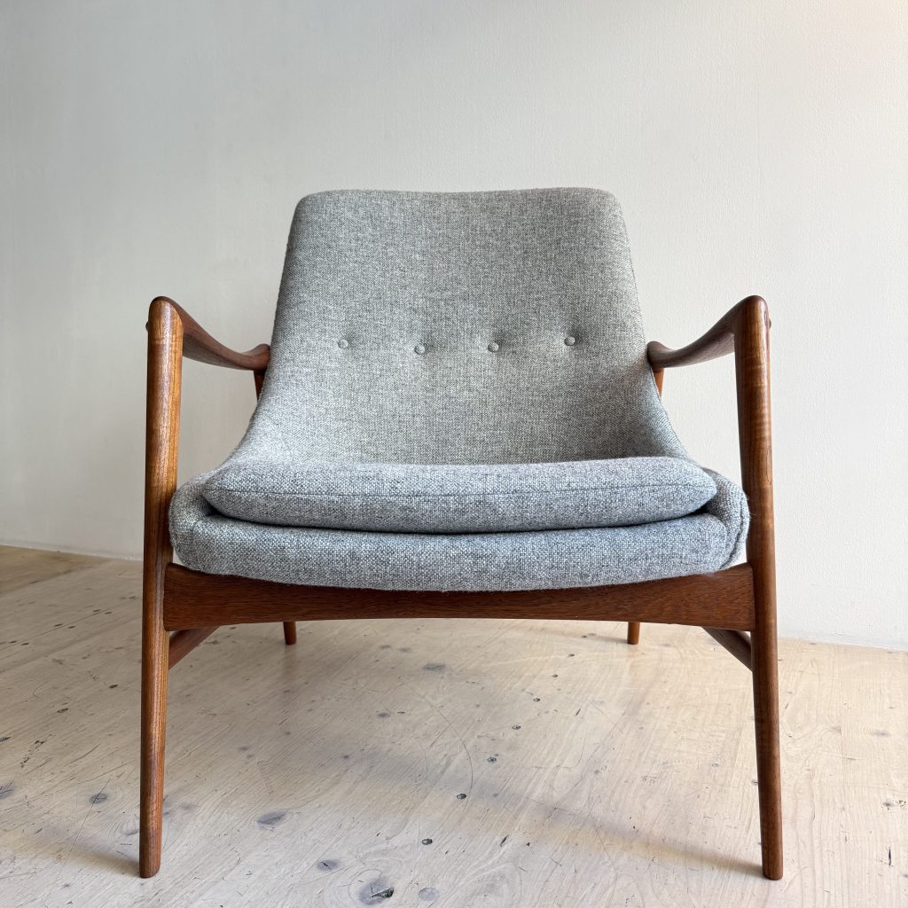 A mid-century modern chair with a gray upholstered seat and backrest, featuring wooden armrests and legs, set against a plain wall.