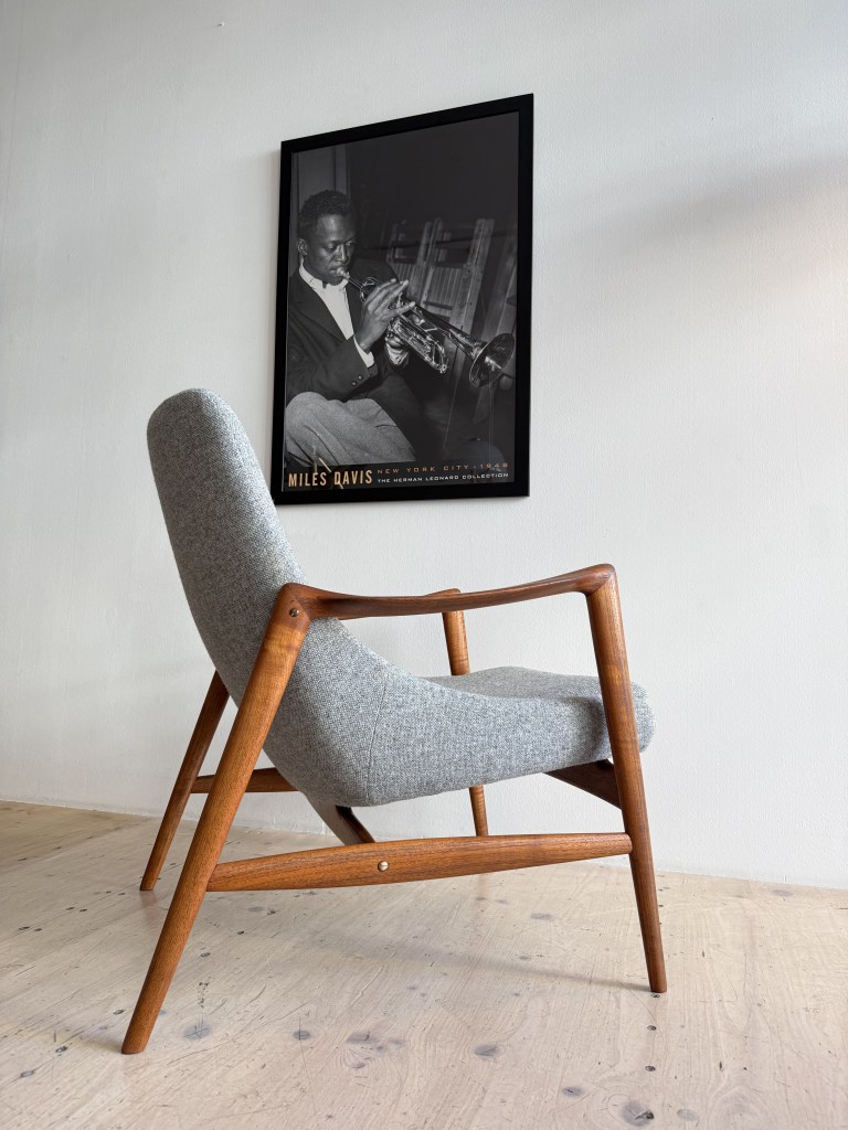 A mid-century modern armchair with a gray fabric seat and wooden legs, positioned next to a black and white framed photo of a musician playing the trumpet.