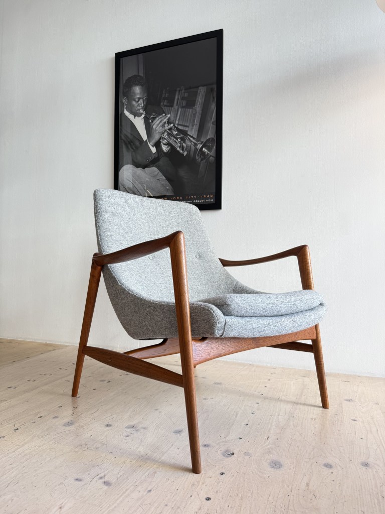 A mid-century modern armchair with a light gray fabric seat and wooden legs, positioned in a stylish room with a black and white framed photograph of a musician playing the trumpet on the wall.