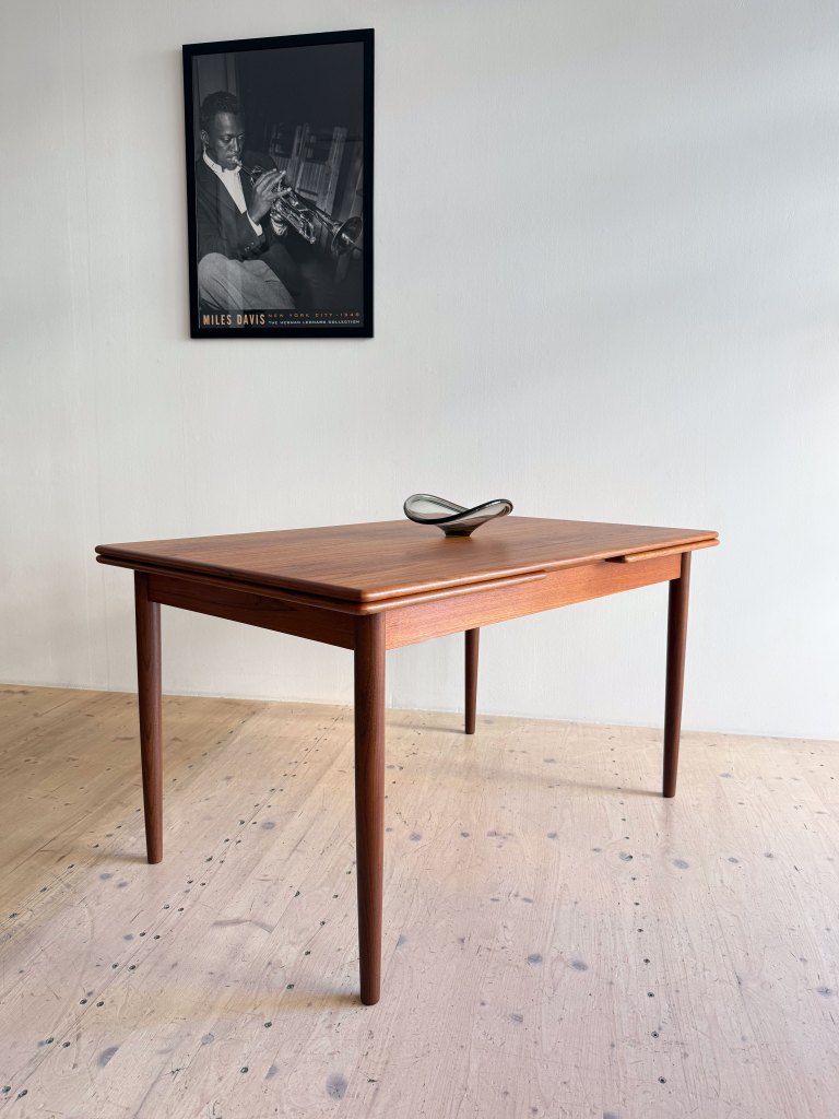 A wooden table with tapered legs and a smooth surface, featuring a decorative object in the center, with a black and white framed poster of a jazz musician on the wall behind it.
