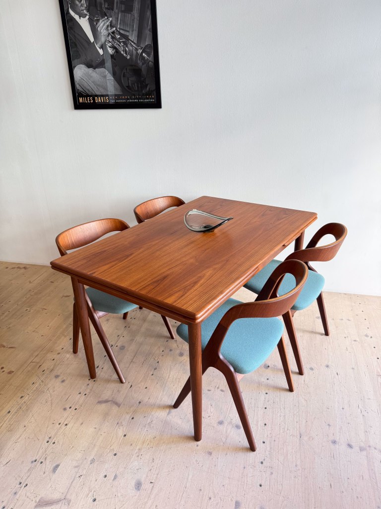 A modern wooden dining table with four light blue upholstered chairs, featuring a sleek design. A decorative glass piece is placed on the table, and a black and white poster of a jazz musician is visible on the wall behind.