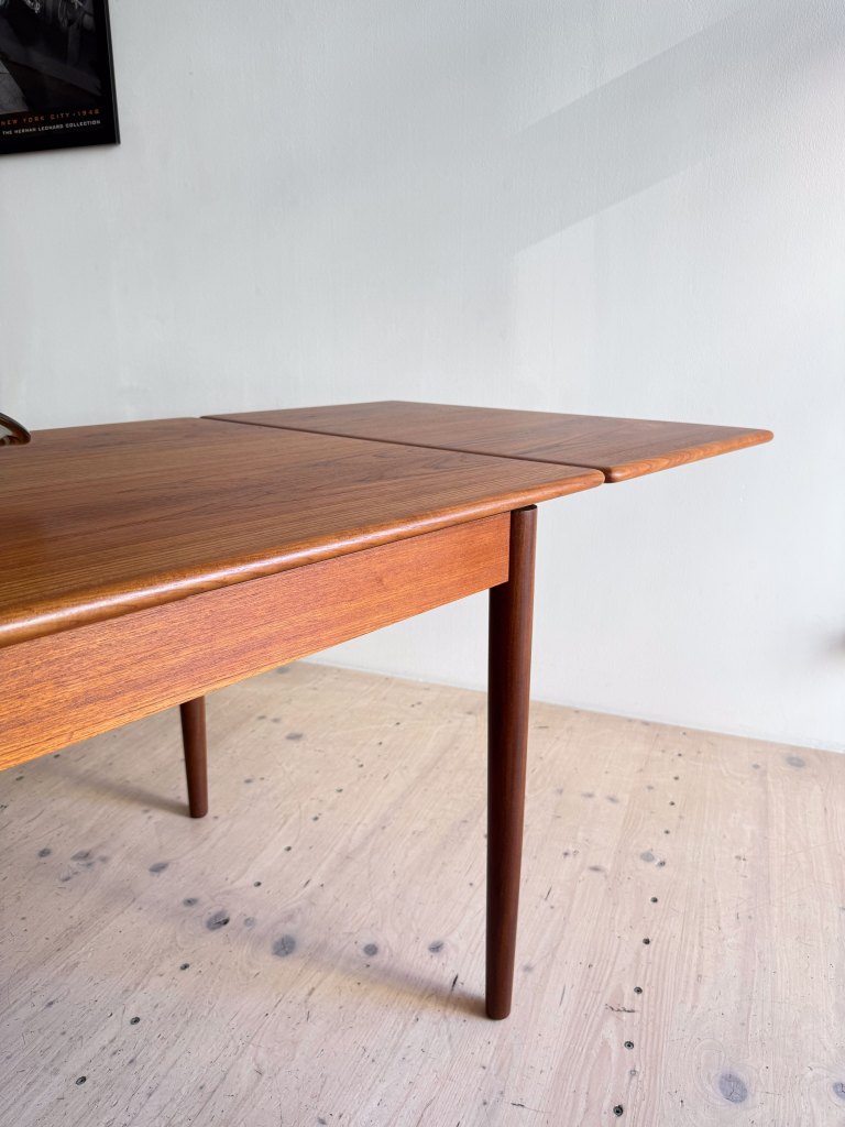 A close-up view of a mid-century modern wooden table with a folding leaf design, featuring tapered legs and a rich brown finish, set against a light-colored wall.