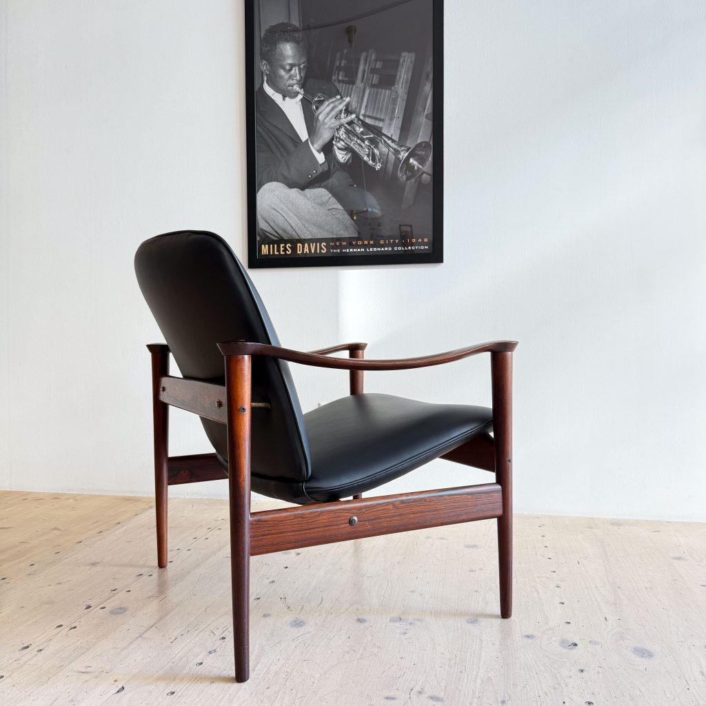 A stylish wooden chair with a black cushion, positioned in front of a white wall featuring a black and white poster of a musician playing the trumpet.